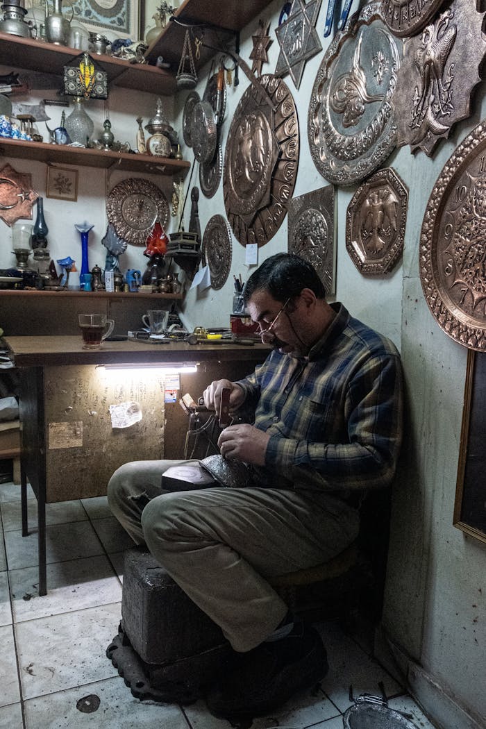 Artisan intricately engraves metal in a cozy workshop filled with ornate wall plaques.