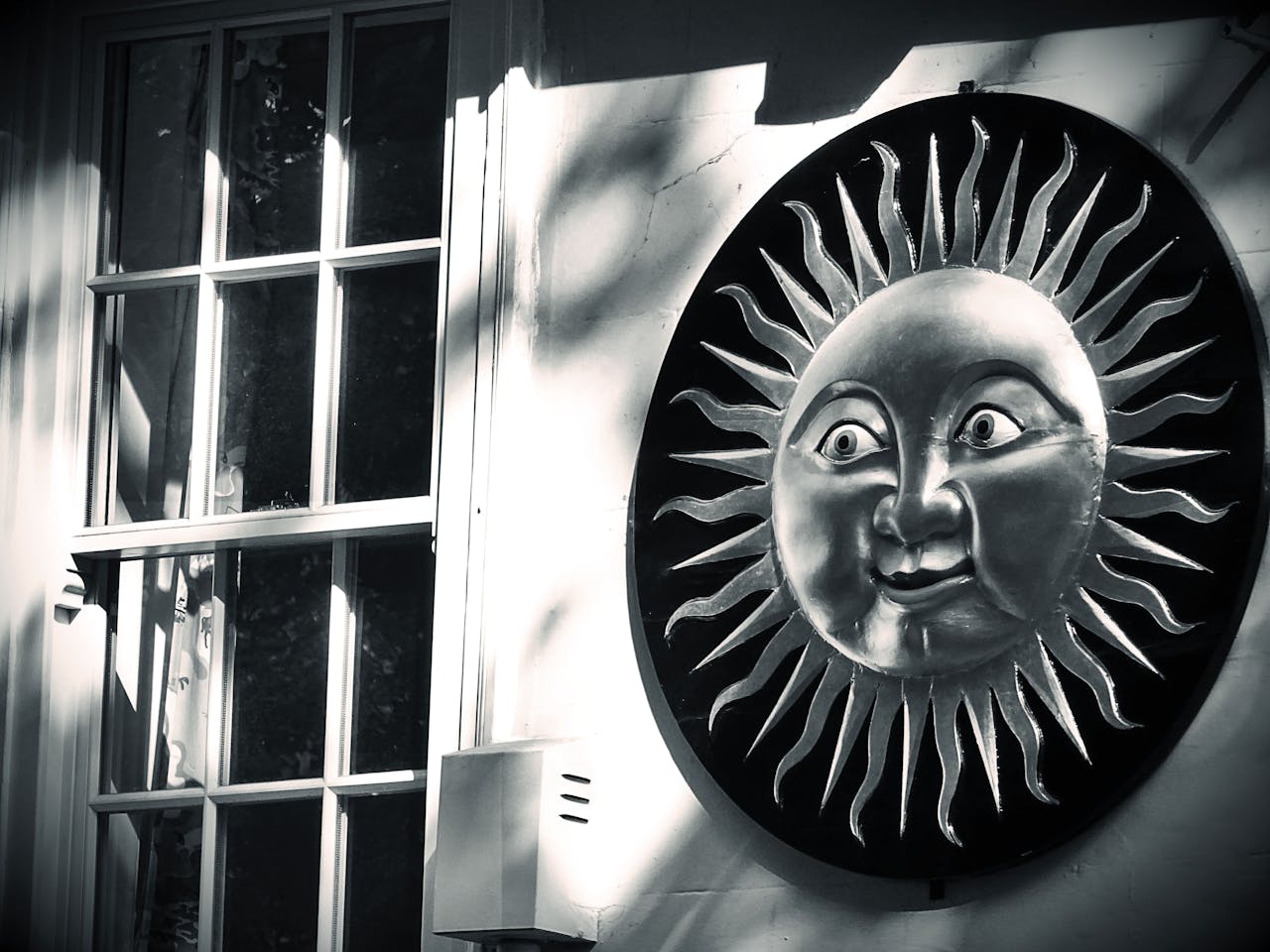 Black and white photo of a metal sun decoration on a wall, adjacent to a window casting shadows.